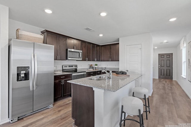 kitchen featuring sink, appliances with stainless steel finishes, a kitchen island with sink, light stone counters, and light wood-type flooring