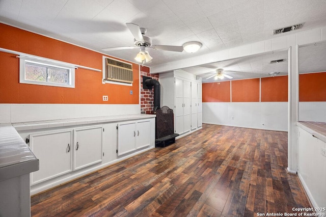 interior space with dark hardwood / wood-style floors, a wall mounted air conditioner, a wood stove, white cabinets, and ceiling fan