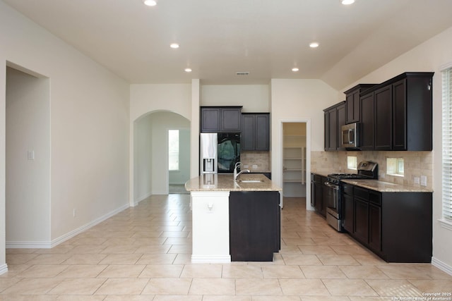 kitchen featuring sink, appliances with stainless steel finishes, backsplash, light stone countertops, and a center island with sink