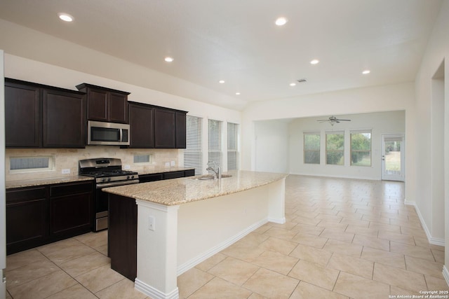 kitchen featuring an island with sink, decorative backsplash, ceiling fan, light stone counters, and stainless steel appliances