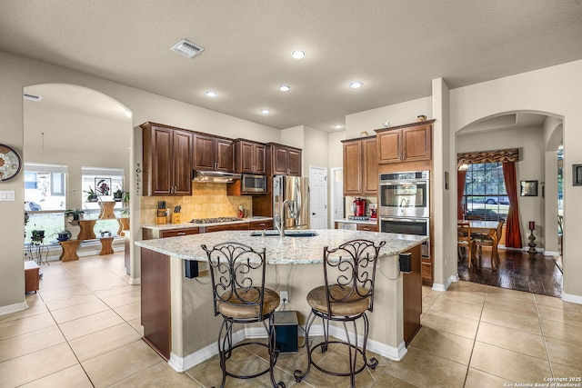 kitchen with appliances with stainless steel finishes, sink, a kitchen breakfast bar, a kitchen island with sink, and light stone countertops