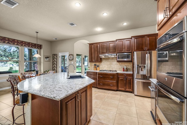 kitchen featuring sink, decorative light fixtures, an island with sink, stainless steel appliances, and light stone countertops