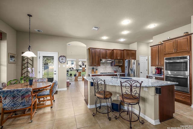 kitchen with appliances with stainless steel finishes, pendant lighting, a kitchen bar, light stone countertops, and a center island with sink