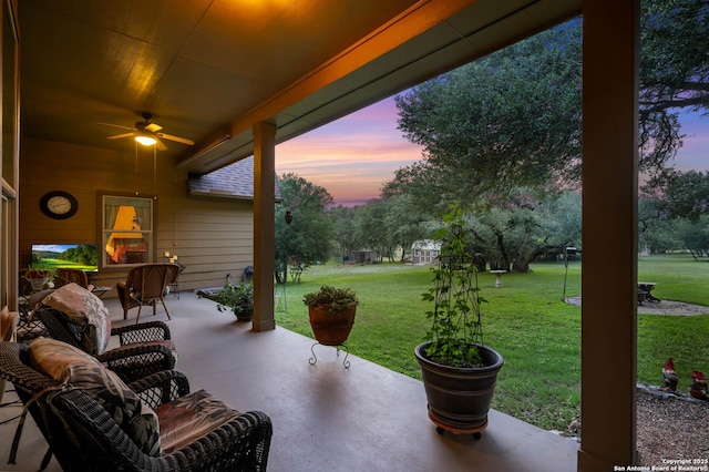 patio terrace at dusk with ceiling fan, a fireplace, and a lawn