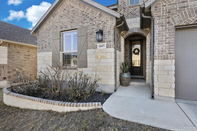 doorway to property featuring brick siding, stone siding, a garage, and roof with shingles