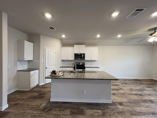 kitchen featuring appliances with stainless steel finishes, white cabinets, dark hardwood / wood-style flooring, dark stone counters, and a center island with sink