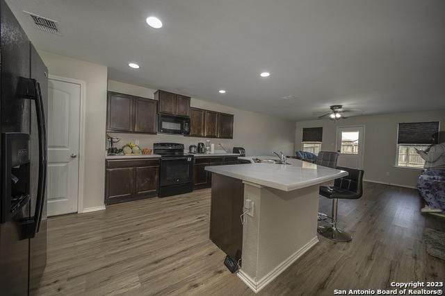 kitchen featuring hardwood / wood-style flooring, a breakfast bar, dark brown cabinetry, black appliances, and a center island with sink