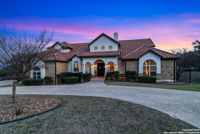 mediterranean / spanish-style house featuring fence, a chimney, curved driveway, stone siding, and a tiled roof