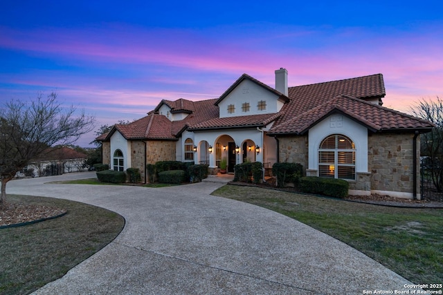 mediterranean / spanish home with stone siding, curved driveway, a tiled roof, and a chimney