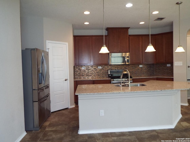 kitchen featuring light stone counters, appliances with stainless steel finishes, decorative light fixtures, and a kitchen island with sink