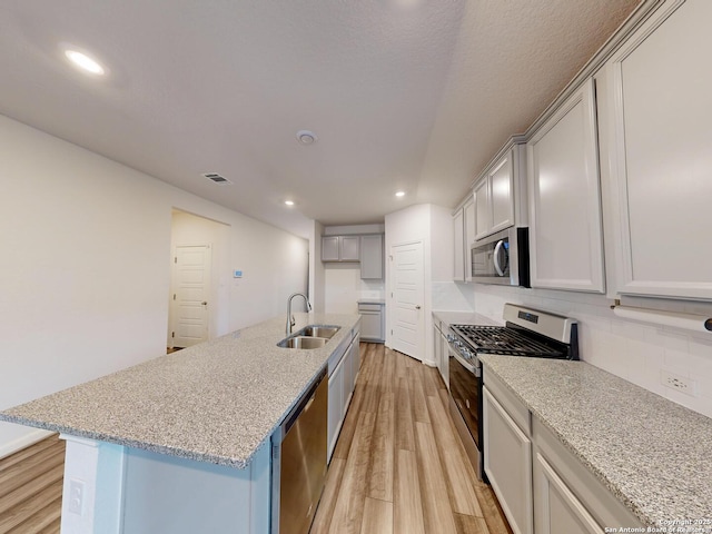 kitchen with sink, a center island with sink, light wood-type flooring, stainless steel appliances, and light stone countertops