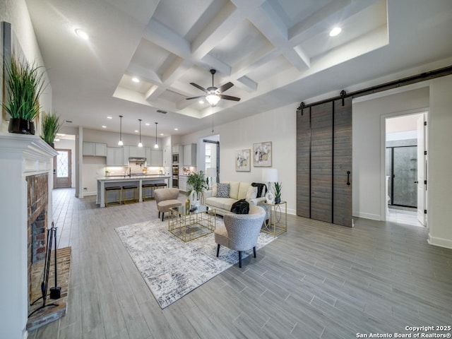 living room with ceiling fan, beam ceiling, coffered ceiling, light hardwood / wood-style floors, and a barn door