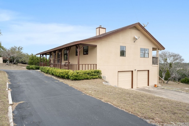 view of side of home featuring a garage, driveway, a chimney, a porch, and stucco siding