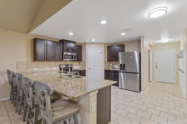 kitchen featuring a breakfast bar area, appliances with stainless steel finishes, dark brown cabinets, light stone countertops, and kitchen peninsula