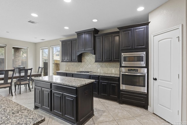 kitchen with light tile patterned floors, stainless steel appliances, light stone countertops, a kitchen island, and decorative backsplash