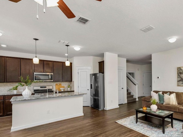 kitchen featuring dark brown cabinetry, light stone counters, hanging light fixtures, appliances with stainless steel finishes, and a kitchen island with sink