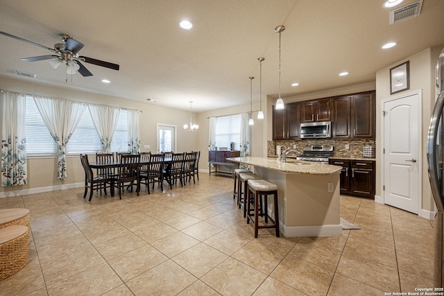 kitchen with hanging light fixtures, stainless steel appliances, dark brown cabinetry, light stone countertops, and a center island with sink