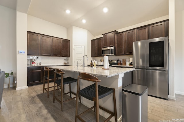 kitchen with a kitchen breakfast bar, dark brown cabinetry, stainless steel appliances, light stone countertops, and a center island with sink