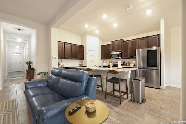 kitchen with a breakfast bar, dark brown cabinets, stainless steel appliances, light stone counters, and an island with sink