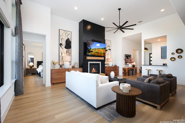 living room with ceiling fan, a large fireplace, a high ceiling, and light wood-type flooring