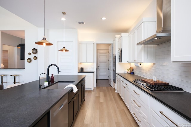 kitchen with appliances with stainless steel finishes, white cabinetry, sink, hanging light fixtures, and wall chimney exhaust hood