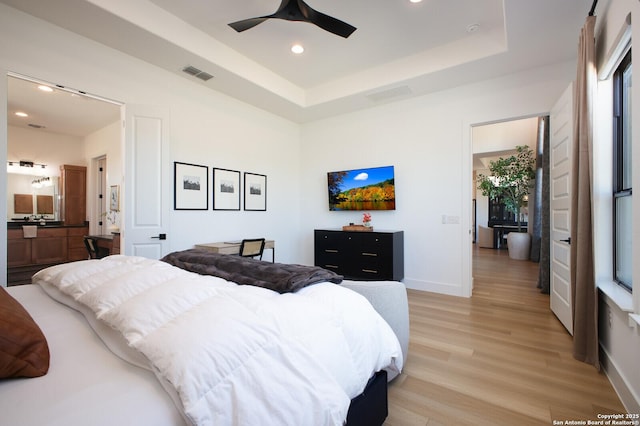 bedroom with ceiling fan, ensuite bathroom, a raised ceiling, and light wood-type flooring