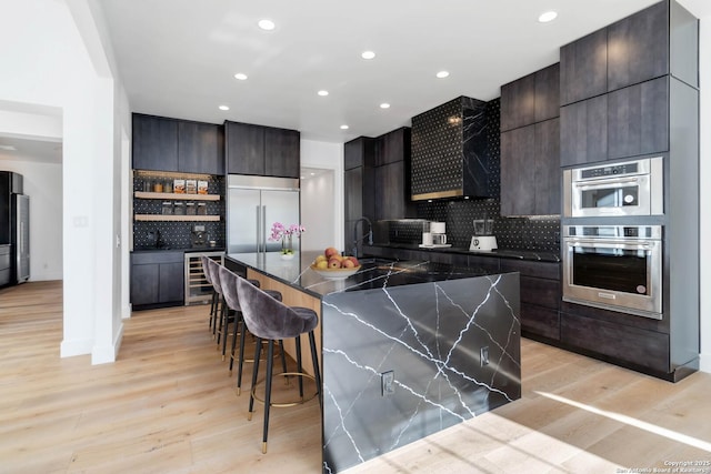 kitchen with a kitchen bar, tasteful backsplash, appliances with stainless steel finishes, an island with sink, and beverage cooler