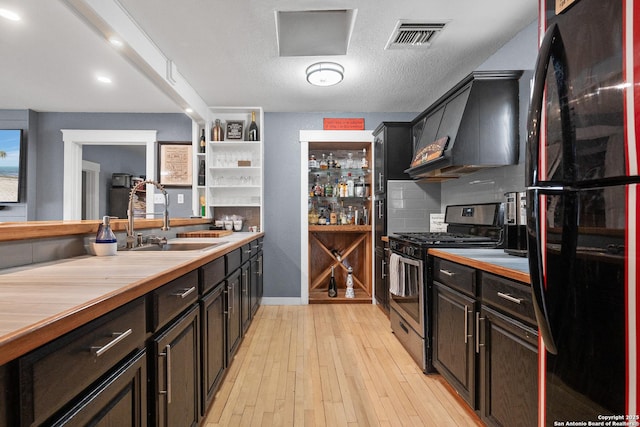 kitchen featuring stainless steel gas range, sink, custom exhaust hood, black fridge, and light wood-type flooring