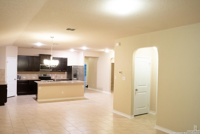 kitchen featuring an island with sink, backsplash, hanging light fixtures, stainless steel appliances, and an inviting chandelier