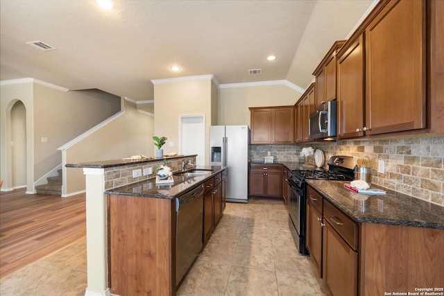 kitchen with sink, a kitchen island with sink, stainless steel appliances, decorative backsplash, and dark stone counters