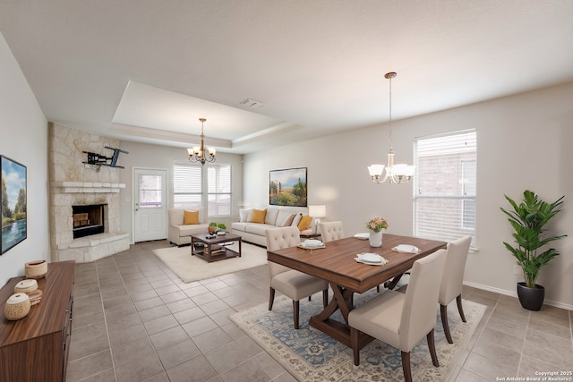 dining space featuring a raised ceiling, light tile patterned flooring, a stone fireplace, and a notable chandelier