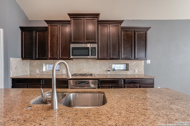 kitchen featuring dark brown cabinetry, decorative backsplash, appliances with stainless steel finishes, light stone counters, and a sink