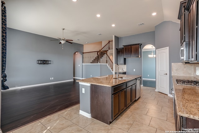 kitchen with arched walkways, a center island with sink, a sink, dark brown cabinets, and light stone countertops