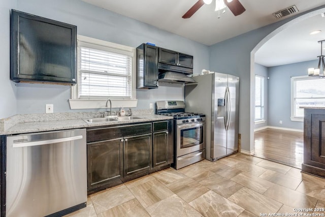 kitchen with sink, light stone counters, hanging light fixtures, stainless steel appliances, and ceiling fan with notable chandelier