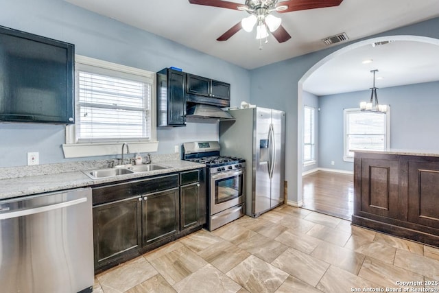 kitchen with appliances with stainless steel finishes, ceiling fan with notable chandelier, sink, hanging light fixtures, and light stone countertops