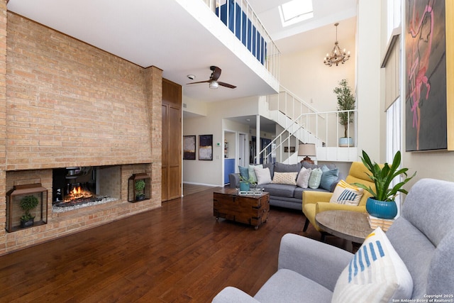 living room with a skylight, a brick fireplace, dark hardwood / wood-style floors, a towering ceiling, and ceiling fan with notable chandelier