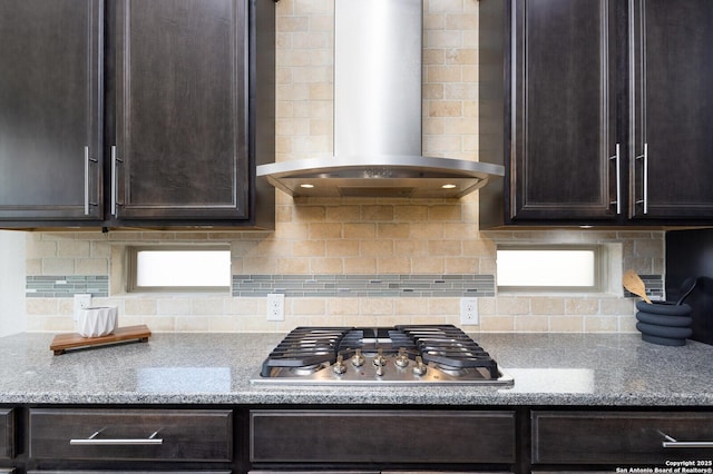 kitchen featuring dark brown cabinetry, wall chimney exhaust hood, light stone counters, tasteful backsplash, and stainless steel gas stovetop