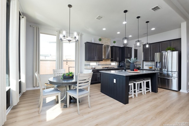 kitchen with a kitchen island with sink, stainless steel fridge with ice dispenser, decorative light fixtures, and wall chimney range hood