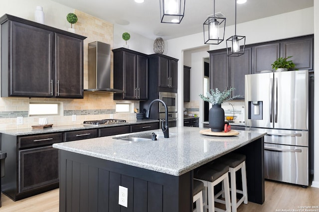 kitchen featuring appliances with stainless steel finishes, sink, a kitchen island with sink, light stone countertops, and wall chimney exhaust hood