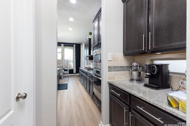 kitchen with dark brown cabinetry, light stone counters, oven, light hardwood / wood-style floors, and backsplash