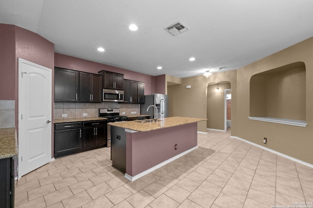 kitchen with light tile patterned floors, a kitchen island with sink, stainless steel appliances, tasteful backsplash, and light stone countertops