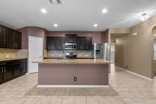 kitchen featuring dark brown cabinets, appliances with stainless steel finishes, light stone countertops, and a center island with sink