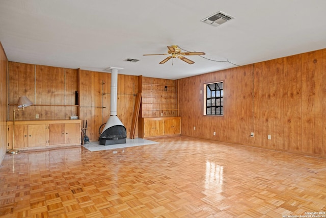 unfurnished living room featuring ceiling fan, light parquet flooring, wooden walls, and a wood stove