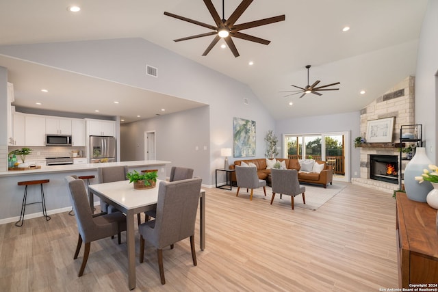 dining area with ceiling fan, a stone fireplace, high vaulted ceiling, and light wood-type flooring