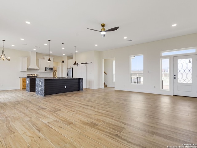 unfurnished living room with a barn door, sink, ceiling fan with notable chandelier, and light hardwood / wood-style floors