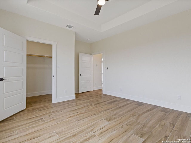 unfurnished bedroom featuring a spacious closet, light hardwood / wood-style flooring, a tray ceiling, a closet, and ceiling fan