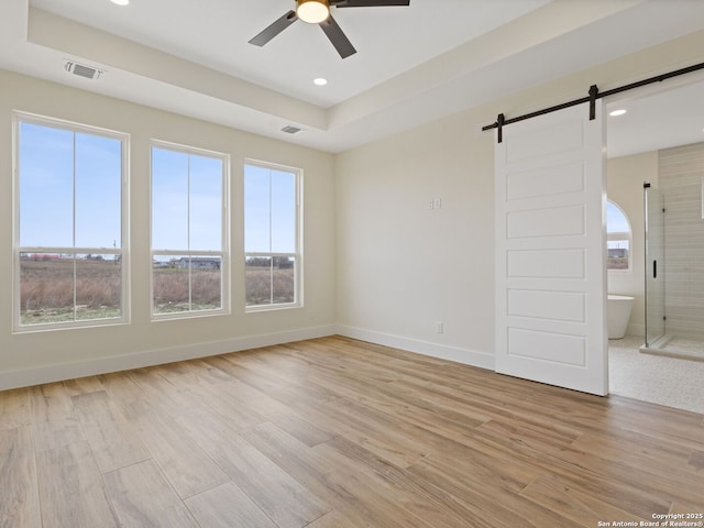 empty room with ceiling fan, a barn door, a raised ceiling, and light hardwood / wood-style floors