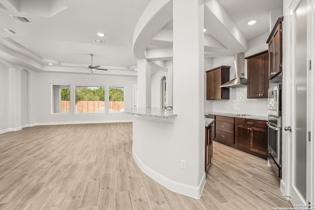 kitchen with light stone counters, stainless steel oven, light wood-type flooring, decorative backsplash, and wall chimney range hood