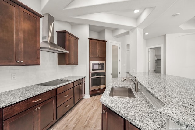 kitchen with sink, black electric stovetop, stainless steel oven, light stone counters, and wall chimney range hood