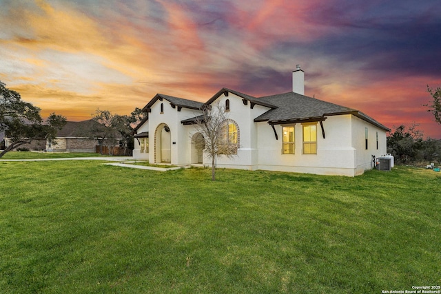 view of front facade with central AC unit, a shingled roof, stucco siding, a front lawn, and a chimney